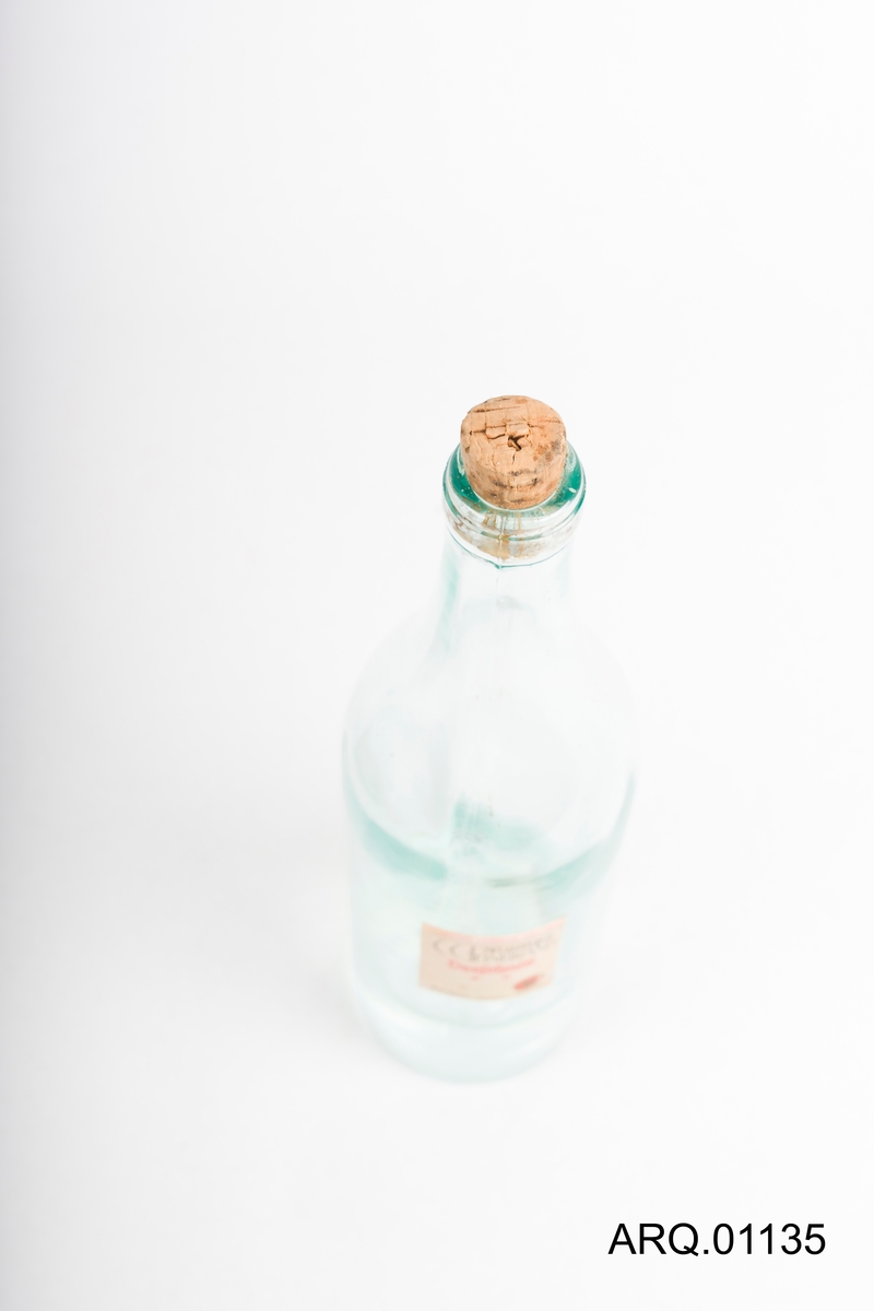 Gjennomsiktig glassflaske med blå-skjær. Liten Etikett med rød og svart skrift. Korken sitter i og det er blankt innhold i flasken.