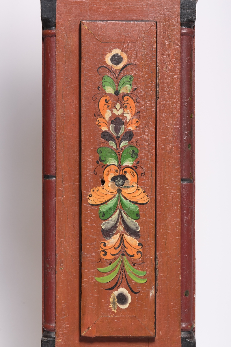 Mørk brunrød, dekorert med påmalte blomster på døra og på toppen. 
(a) kasse
(b) topp/ hode
(c) urverk  - urskive av metall med romerske tall. 
(d) pendel 
(e og f) lodd
(g) sveiv
(h) øvre pyntelist
(i) sokkel
