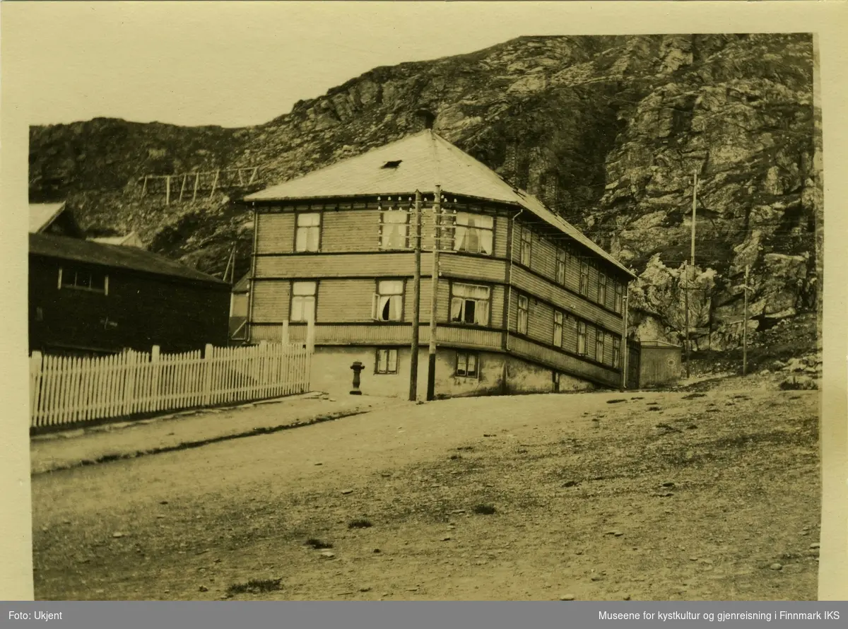 Hammerfest gamlehjem er i 1924 et vakkert trebygg med skifertak som står foran en fjellside med skredsikring. En strømmast og en brannhydrant står foran huset. Til høyre ligger et andre hus med gjerdet rundt.