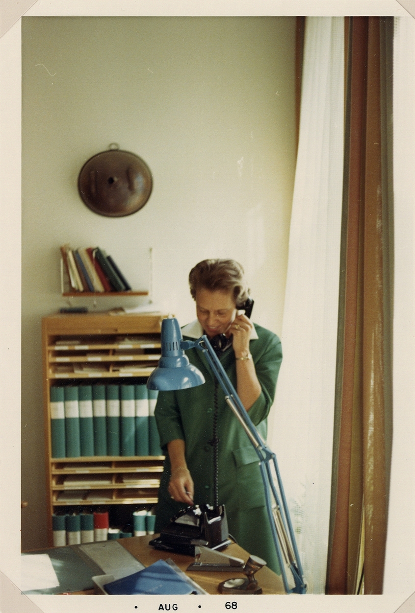 En kvinna i grön skyddsrock pratar i telefon. Färgfoto, augusti 1968. 
Eivor Green, anställd vid Systembolaget, Växjö.