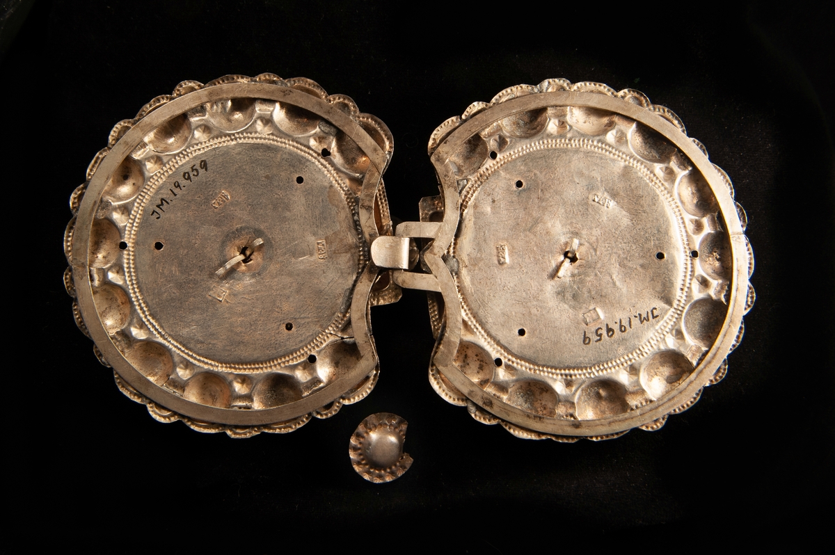 Ett stort tröjspänne av ciselerat silver med pålagda "klippta" ornament i upphöjt arbete (bladform) och en blomma i mitten. Behängd med 3 delvis ciselerade små, runda plattor av silver. Har en hyska som fästes i haken på JM.19959:2.
