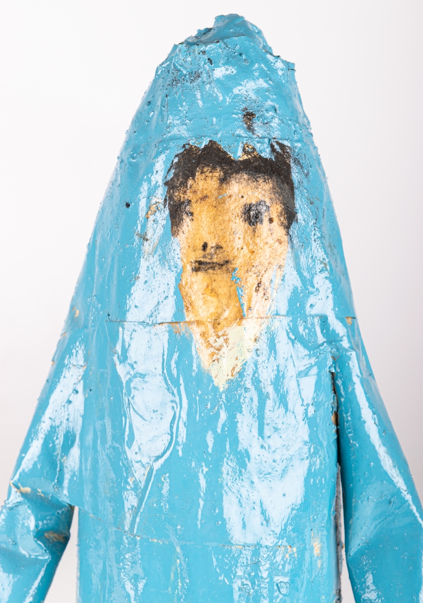 Skulptur av trä och papper. Figur/människofigur, helt blåmålad. Ansikte i beige med detaljer i svart. Mössliknande form på huvudet, med garntofs bak.