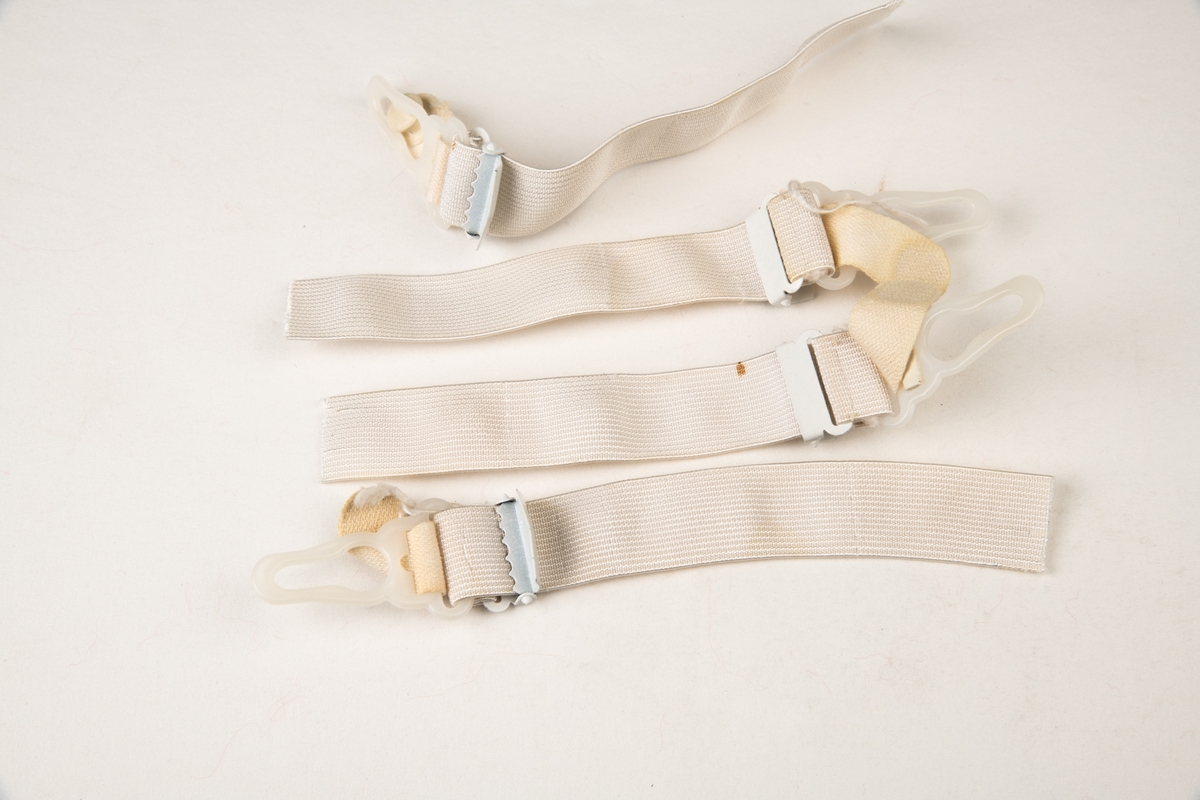 Et sett med fire hvite stropper med en metallspenne og hekter av plast i enden. Selve stroppen er elastisk.