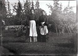 To kvinner i lange kjoler med forklær over står i hage med f