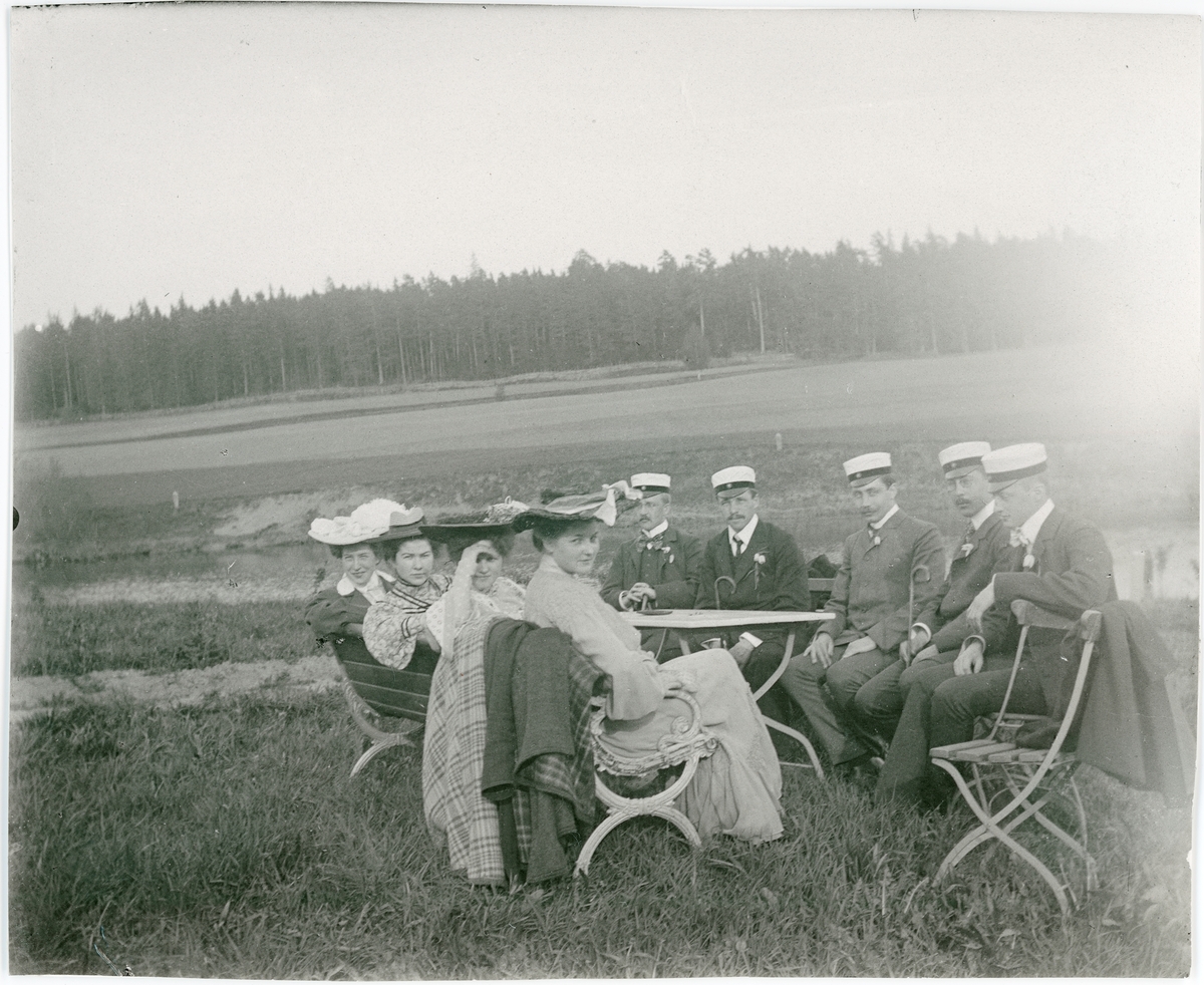 Studenter, kvinnor och män sittande vid bord ute på gräs, skog i bakgrunden, sannolikt Uppland omkring 1900