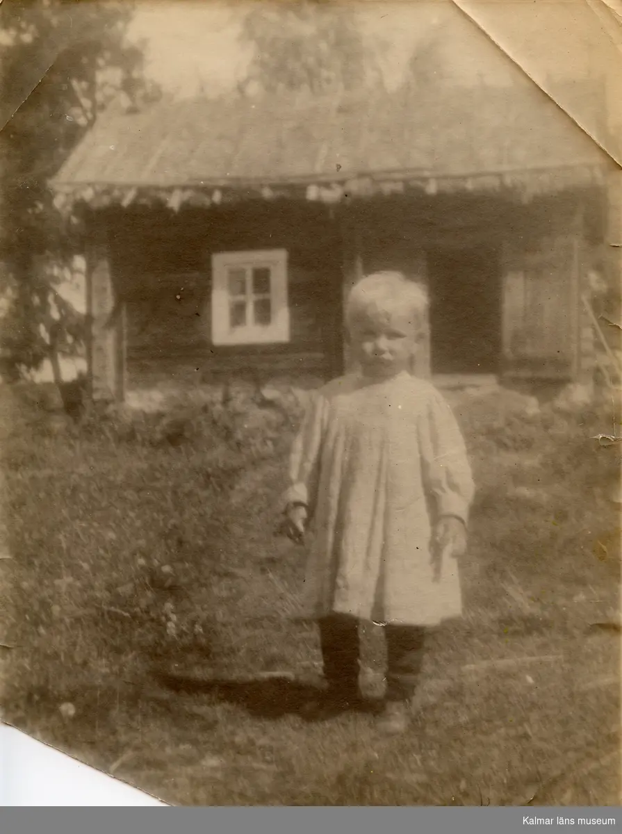 Fotografi av en liten pojke, Curt Nyström Stoopendaa,l klädd i kolt. Han står utanför en liten stuga. Samma negativ som KLM044610-00118.
