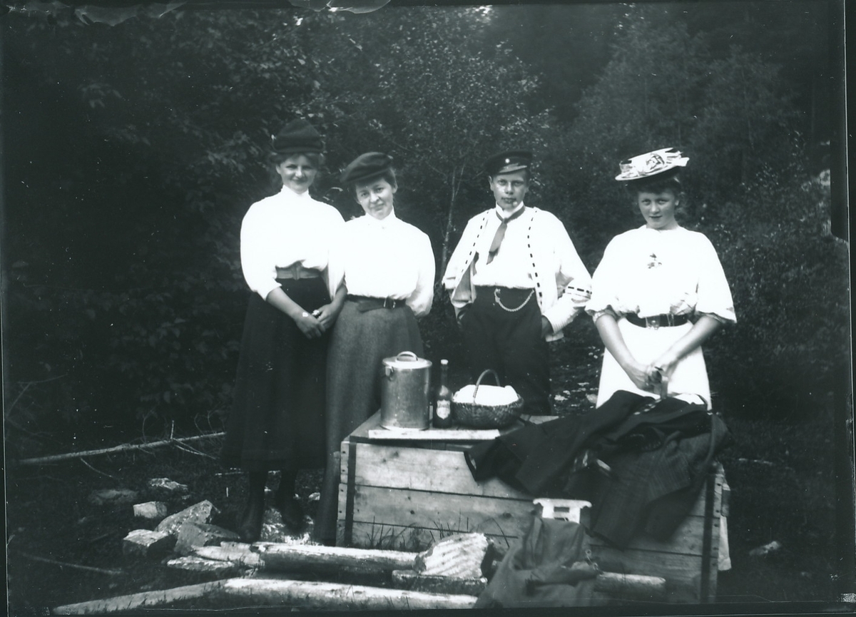 Tre kvinner og en ung mann står bak kasse med spann og kurv oppå. Landtur i skogsterreng.