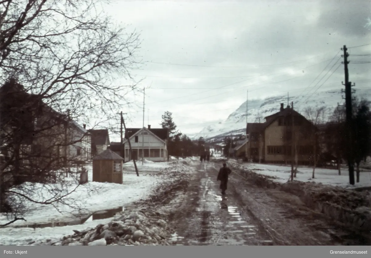 En person spaserer langs en slapsete vei som går mellom flere bygninger. Til venstre står en liten kiosk. Snøen ligger på siden av veien og på fjellene som kan sees i bakgrunnen.