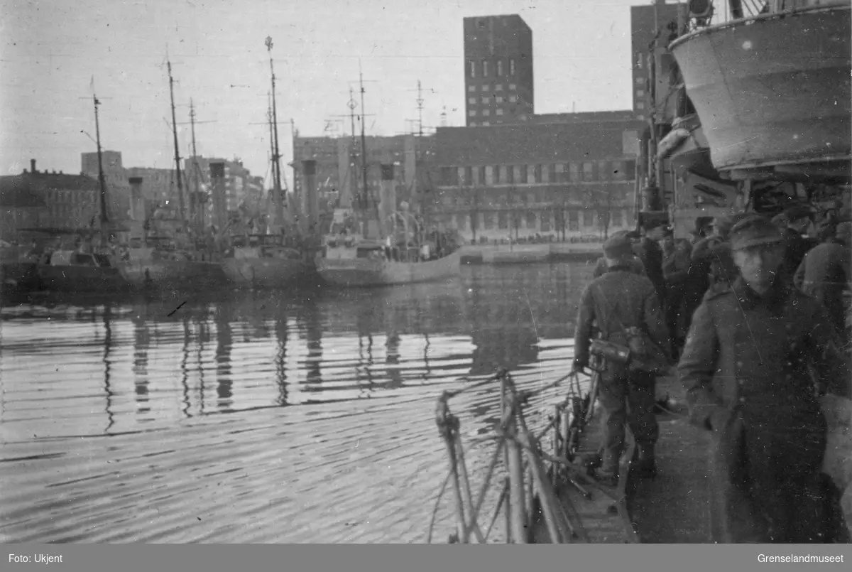 Motiv fra indre havn i Oslo. Bildet er tatt fra et krigsskip der en kan se soldater med uniform og utstyr bevege seg langs rekka. I bakgrunnen ligger Oslo Rådhus. Til venstre ligger minst 4 andre båter fortøyd. 