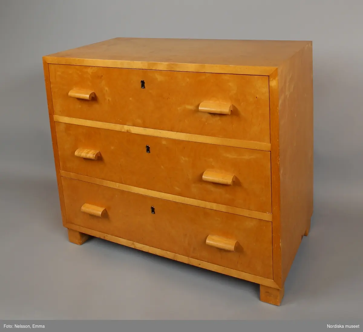 Byrå (A) med tre lådor (B:1-3),"funkis", 1930-40-tal, av ljust polerat trä, sannolikt björk. Rektangulär form på fyra klossformade fötter. Rygg av plywood, spikad. Draghandtag av trä. Instickslås. Nyckelbussningar av metall.
Anm: Lådorna försedda med röd- och vitmönstrat hyllpapper, fäst med häftstift.
/Anna Arfvidsson Womack 2021-08-16