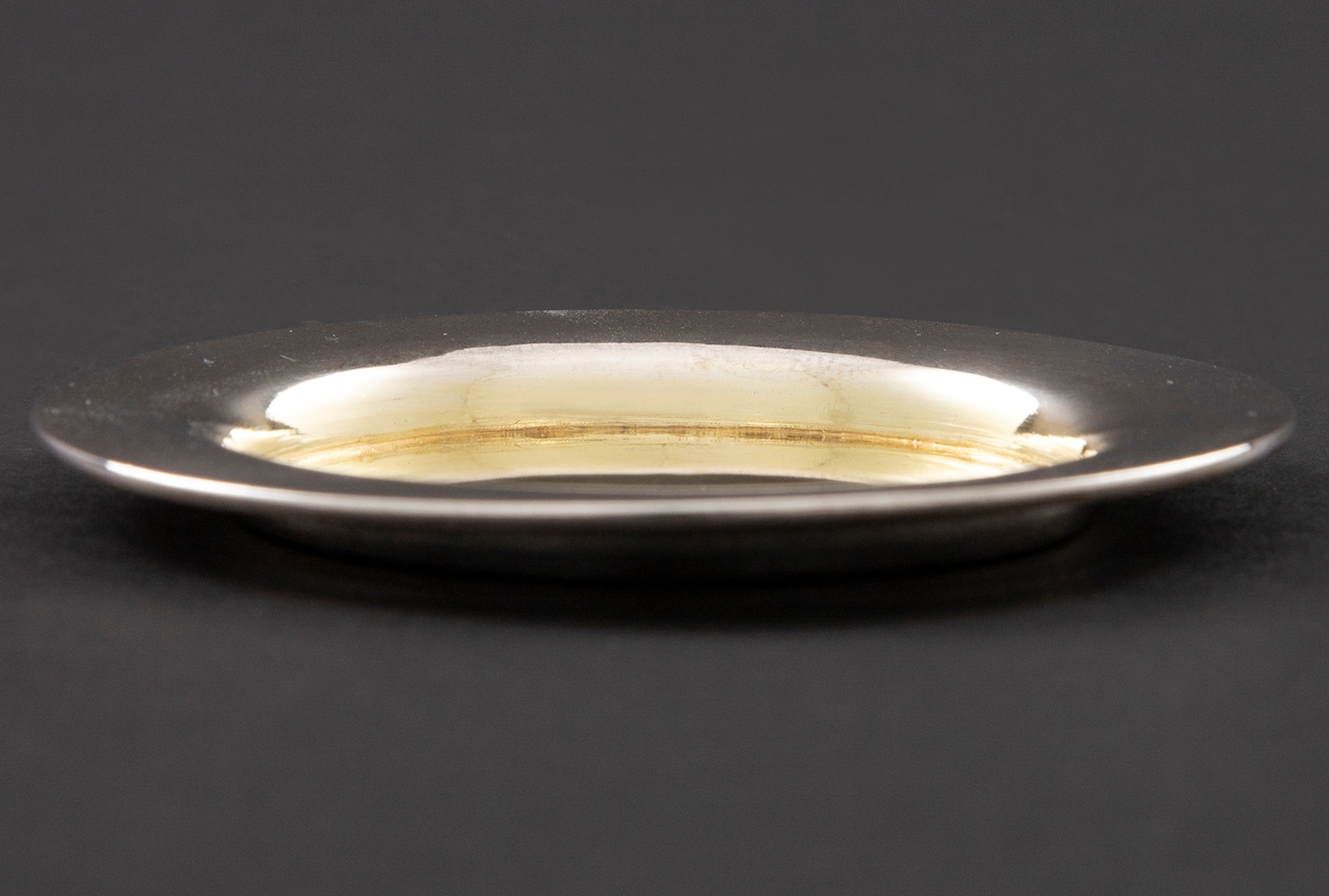 Sirkulær udekorert disk i sølv. Den har forholdsvis bred kant og forgylt midtparti. Formen er rund med rette kanter og svakt hvelvet bunn.