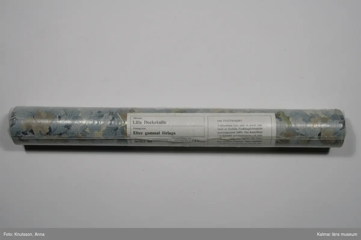 KLM 43780 Tapet i papper. 4 rullar, varav två ligger kvar i originalförpackning. Tapet med vit botten och textilimiterat mönster. Blad och blommor i blått och rosa och grenar i grönt. Mönstret heter Lilla Dockekulla och tapeten är tryckt efter gammal förlaga. Artikelnummer: 30062-01.