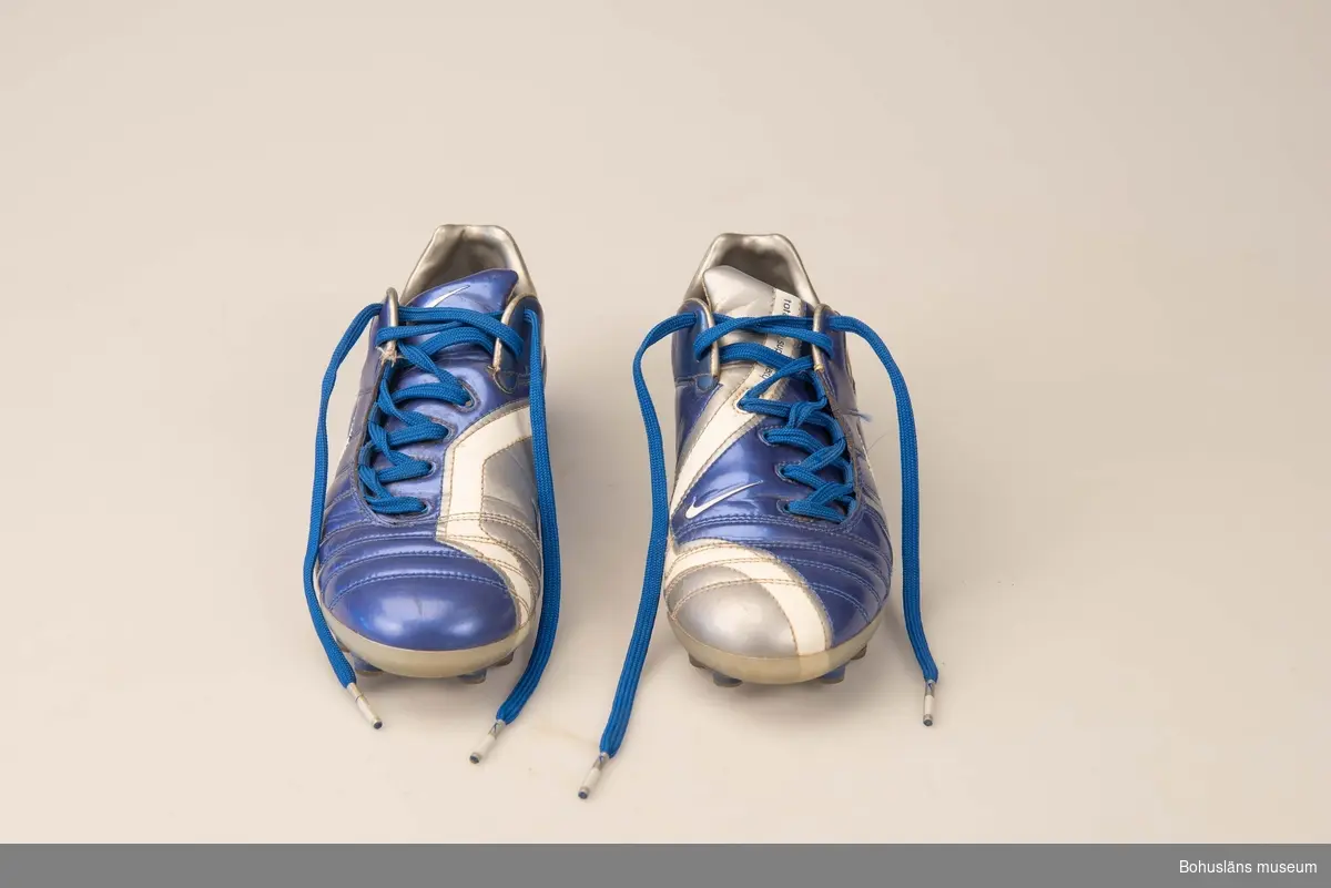 Blå- och vita fotbollskor av amerikanska märket Nike, modell Zoomair 90. Helt i plast, ovanlädret i blank, delvis fodrad mjukplast i blåmetallic, silver och vitt, plösens undersida av konstläder.  Nikes symbol i vitt på tre ställen. Sula med dobbar, åtta undertill fram och fyra underill på hälen. Snörknytning med blått skonöre. På utsidan hälen ingjuten text ZOOMAIR. På plösen texten: 
"total 90 sumpremacy", under plösen papperetikett med texten MADE I N CHINA och storleksangivelse 38.
