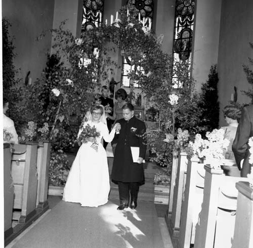 Ett brudpar står i en altargång under en blomsterportal. De håller varandra i händerna.