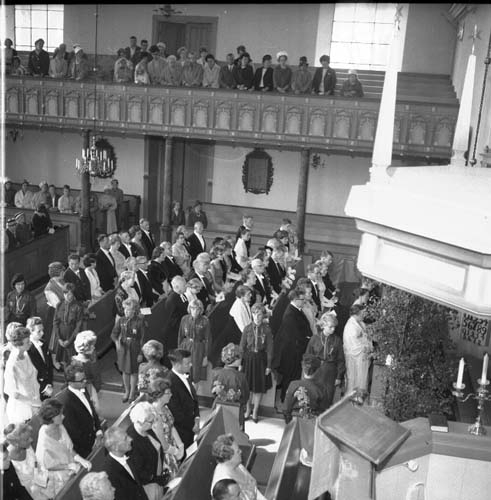 Fotot är taget från läktaren ned mot bröllopsgästerna. I förgrunden syns predikstolen. Alla besökare står upp. Längst med altargången står unga kvinnor uppställda på båda sidorna. De har alla likandnde klänningar.