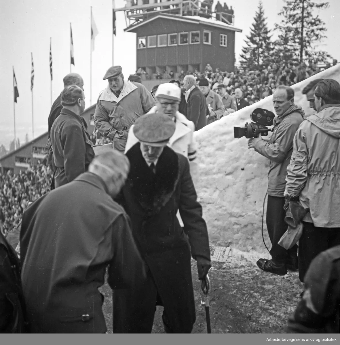 De sjette olympiske vinterleker, Oslo 14.-25. februar 1952. Kong Haakon VII, president i den internasjonale olympiske komité, Avery Brundage og Kronprins Olav på tribunen i Holmenkollen. Til høyre med filmkamera: Finn Bergan.