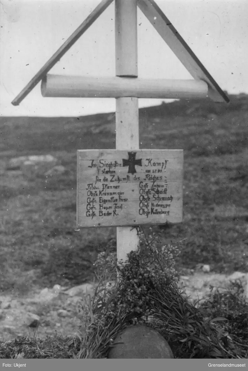 Et kors med en hjelm og blomsterbuketter inntil, står ved et gravsted. På korset er det slått opp en plate med hakekors og noe informasjon med en dato i 1941. Under er det listet opp grad og navn på 10 personer. 