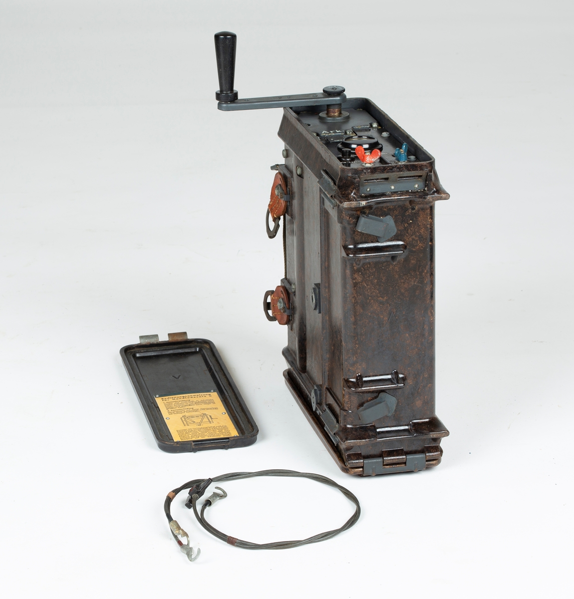 Transportabel generator med sveiv, to kabler. Tyskprodusert, antagelig fra 2. verdenskrig. Til radio og telefonsamband. Tysk bruksanvisning på sidelokk.