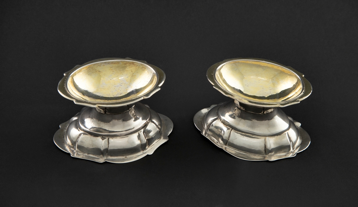To identiske saltkar i sølv. De har oval skål med rett, utstående kant og knekkbuet profil. Skålene står på oval, høyt hvelvet fot med rette knekk. De har rett fotkant med knekkbuet profil. Innvendig er skålene forgylte.