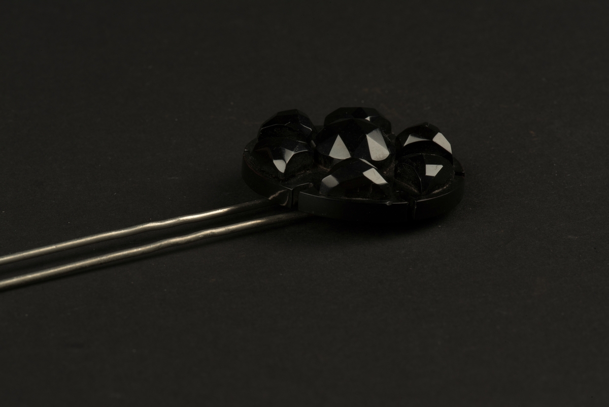 Två hårnålar med rund dekoration i svart.
Nålarna består av parallella långa hårnålar samt en dekoration i form av en mönsterslipad svart platta. Denna är troligen tillverkad i plast, eventuellt cellulosaacetat.