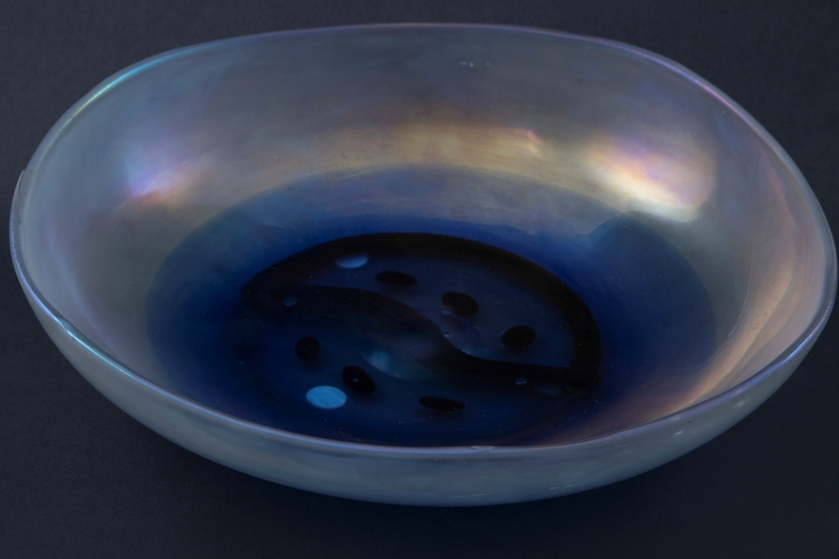 Lav sirkulær glasskål i irisert glass. I bunnen av skålen et blåfarget parti med et sirkulært ornament i brunt og grønt samt overfang.