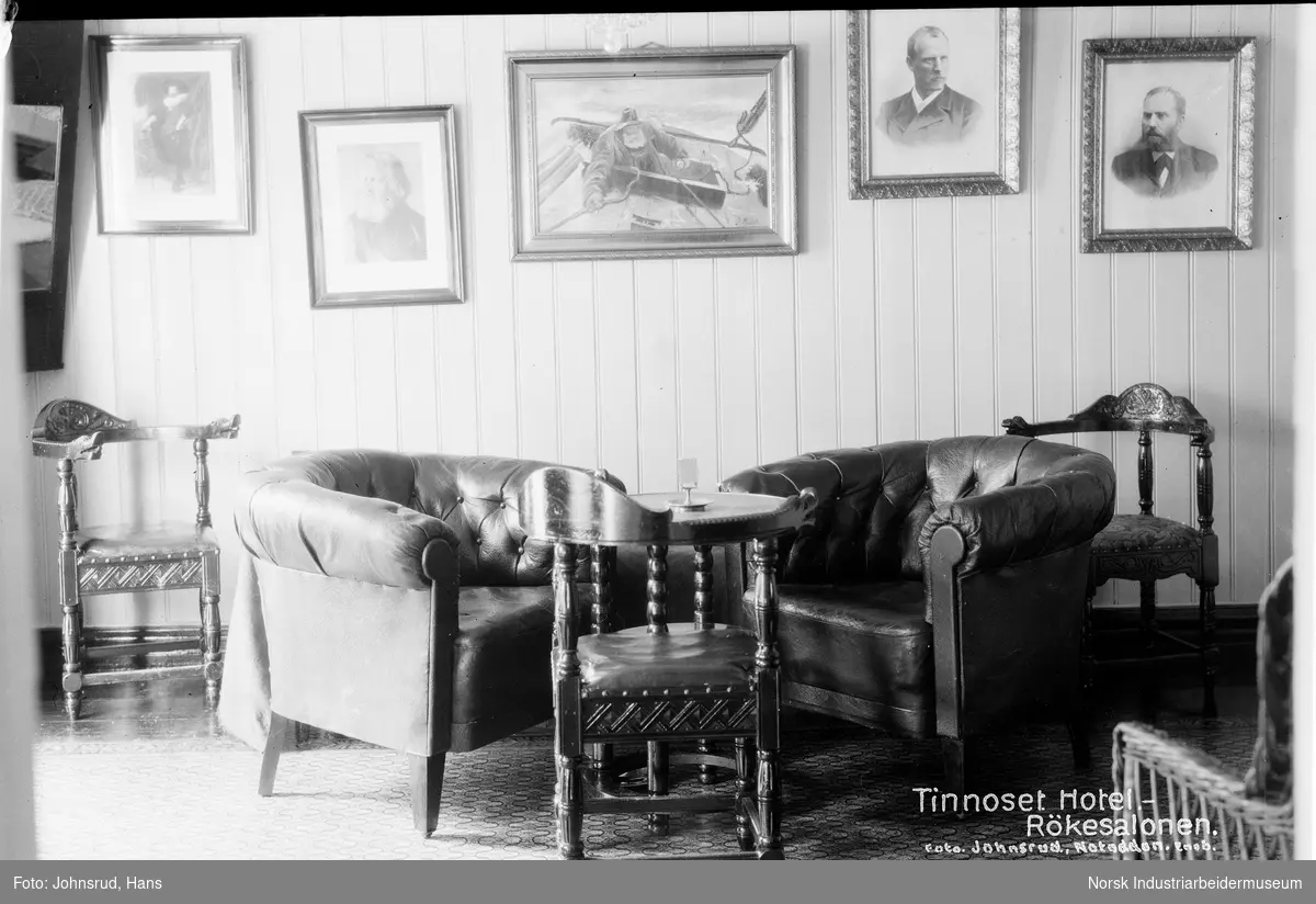 Interiørbilde av røkesalongen ved Tinnoset hotell. Midt i bildet er det tre stoler rundt en bord. Det er to stoler til, en ytterst på hver side av bildet. På veggen i bakgrunnen er det fem bilder.
