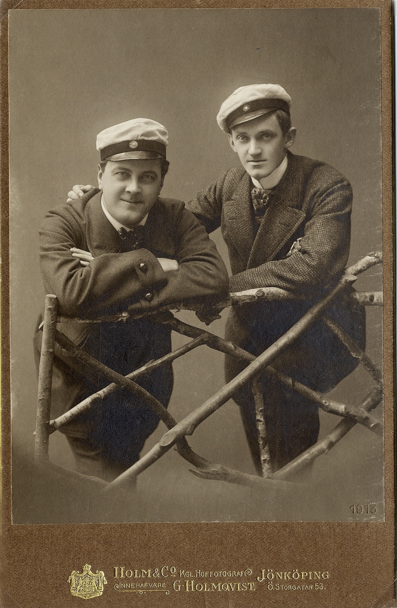 Två män klädda i kostym och studentmössa, lutar sig mot en "gärdesgård".
Knäbild, en face. Ateljéfoto 1902.