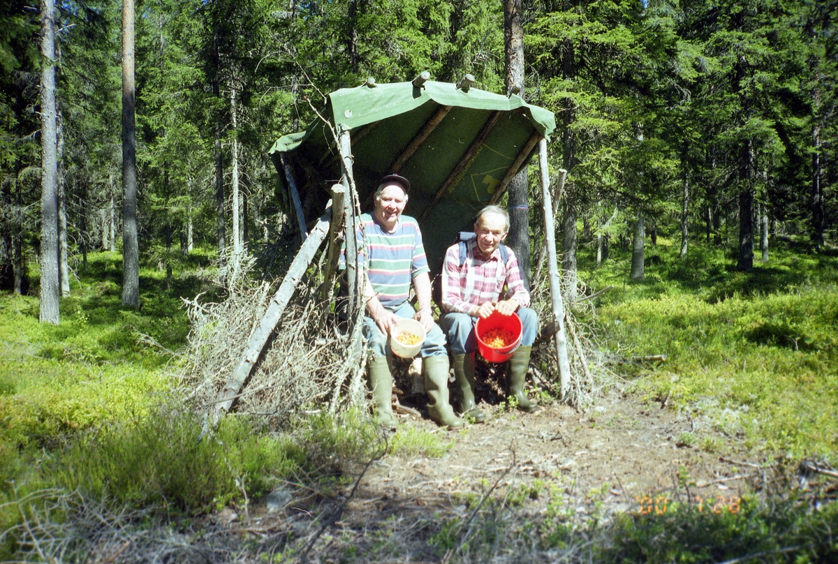 I ett vindskydd i en solig skogsglänta sitter två män. De ler mot fotografen och visar fram varsin hink med hjortron, 28 juli 2000.