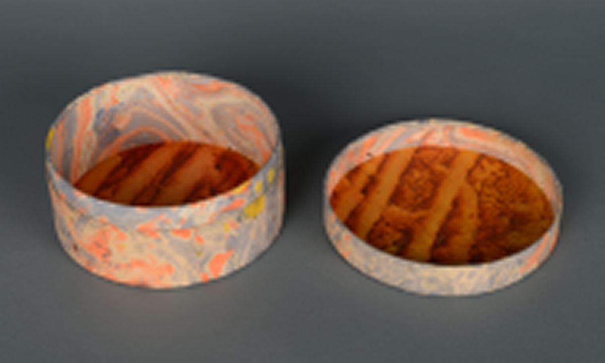 En rund, sylinderformet boks med lokk. Den er laget av papp, og trekt med papir. På utsiden er den grå, med gule og oransje prikker. I bunnen på innsiden og inne i lokket er den oransje med rødt marmorert mønster. På undersiden er den brun. Laget i pappsløyd.
