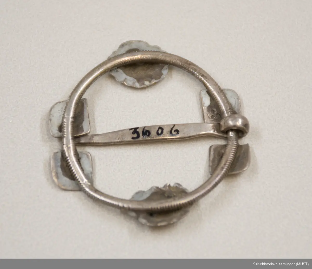 Ringen er av snodd sølv og  en nål er festet på tvers. På ringen er det fire plater og to blomsterornamenter. 