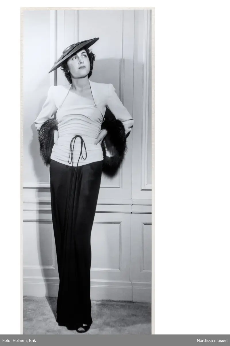 Mannekäng visar festklänning i svart och vitt med rynkat liv med tunn snodd i midjan. Matchande bolero  med pälsdetaljer. Hatt. NK:s Franska damskrädderivåren 1938.