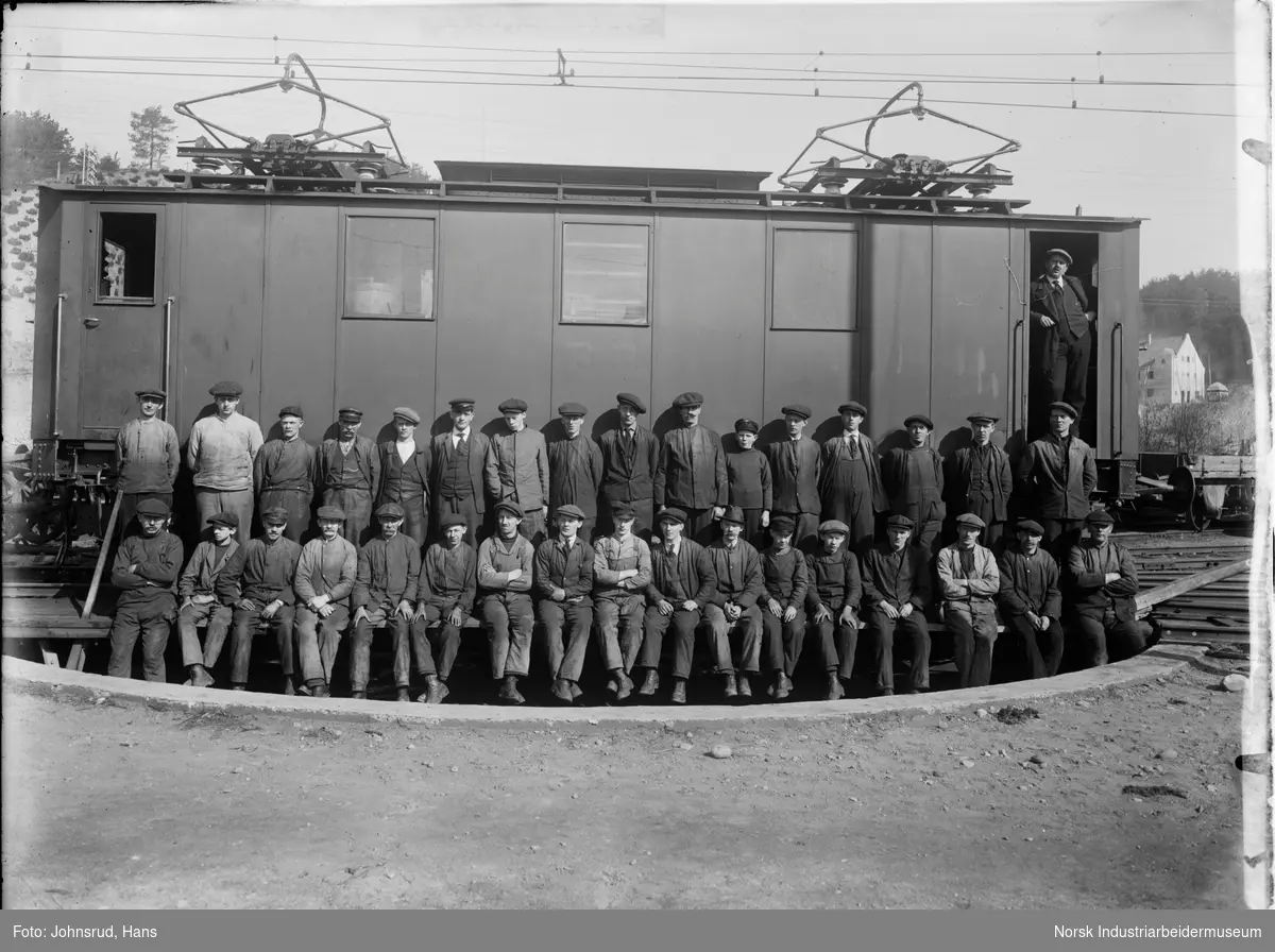 En gruppe jernbanearbeidere fotografert i en svingskive foran et elektrisk lokmotiv ved Rjukanbanen Jernbaneverksted. Lokomotivet er enten RjB 1, 2, eller 3.