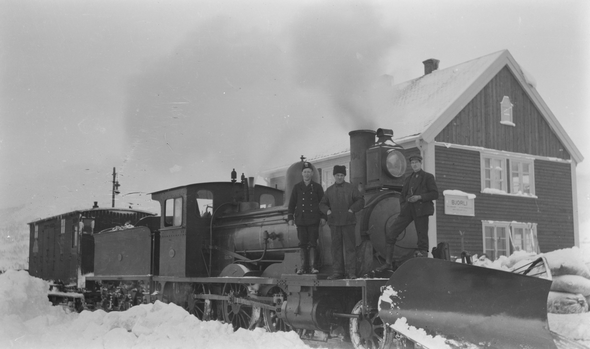 Damplokomotiv Type 15 på Bjorli stasjon med snøplog og en vogn. På lokomotivet står tre menn hvor mannen i uniform antas å være banevokter eller stasjonsmester, og de to andre førere av lokomotivet