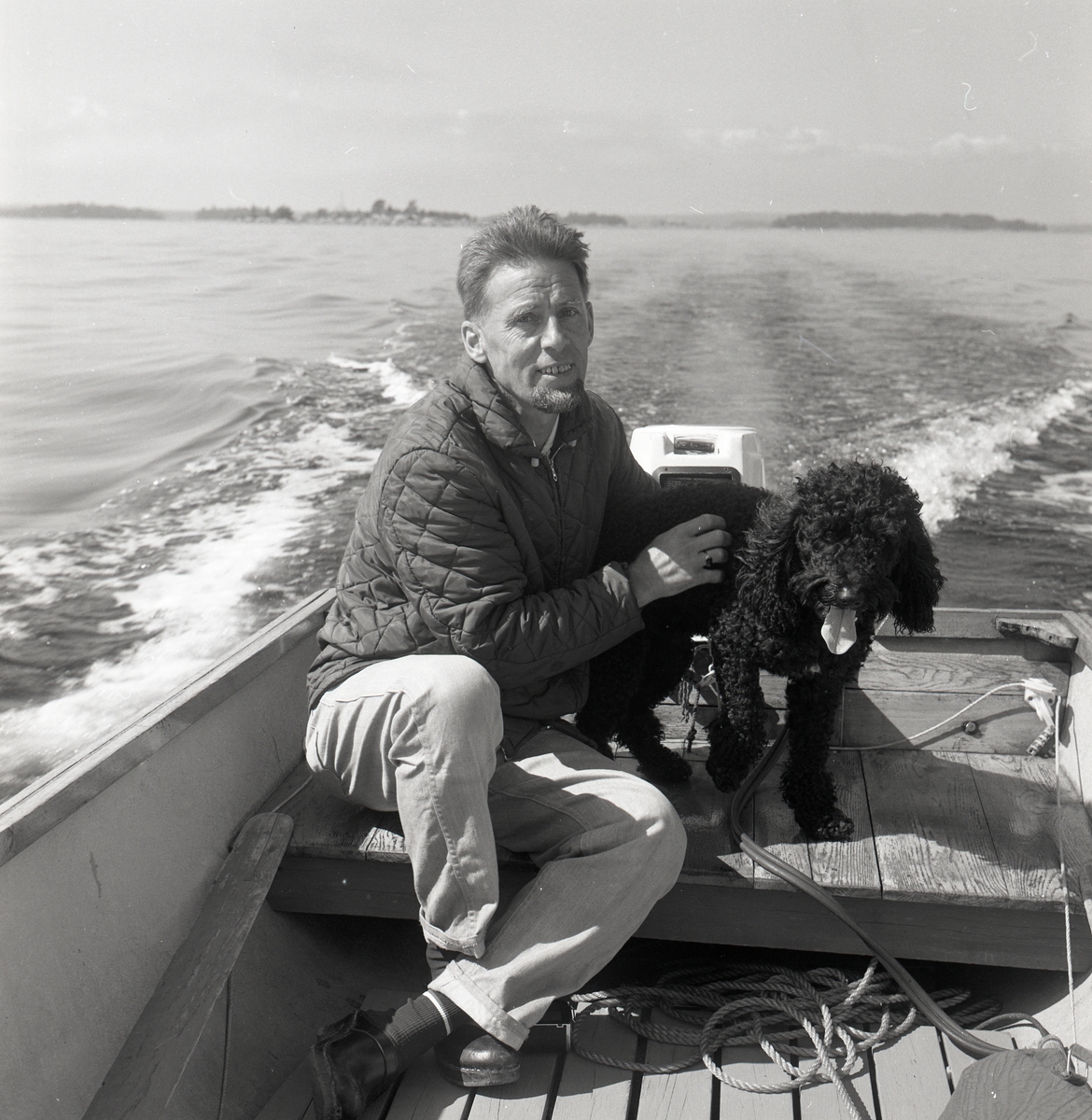 En man sitter vid rodret och kör en motorbåt. Han var mästerlots på Lilljungfruns lotsplats. Intill honom sitter hunden Fabian. Rönnskär, augusti 1969.