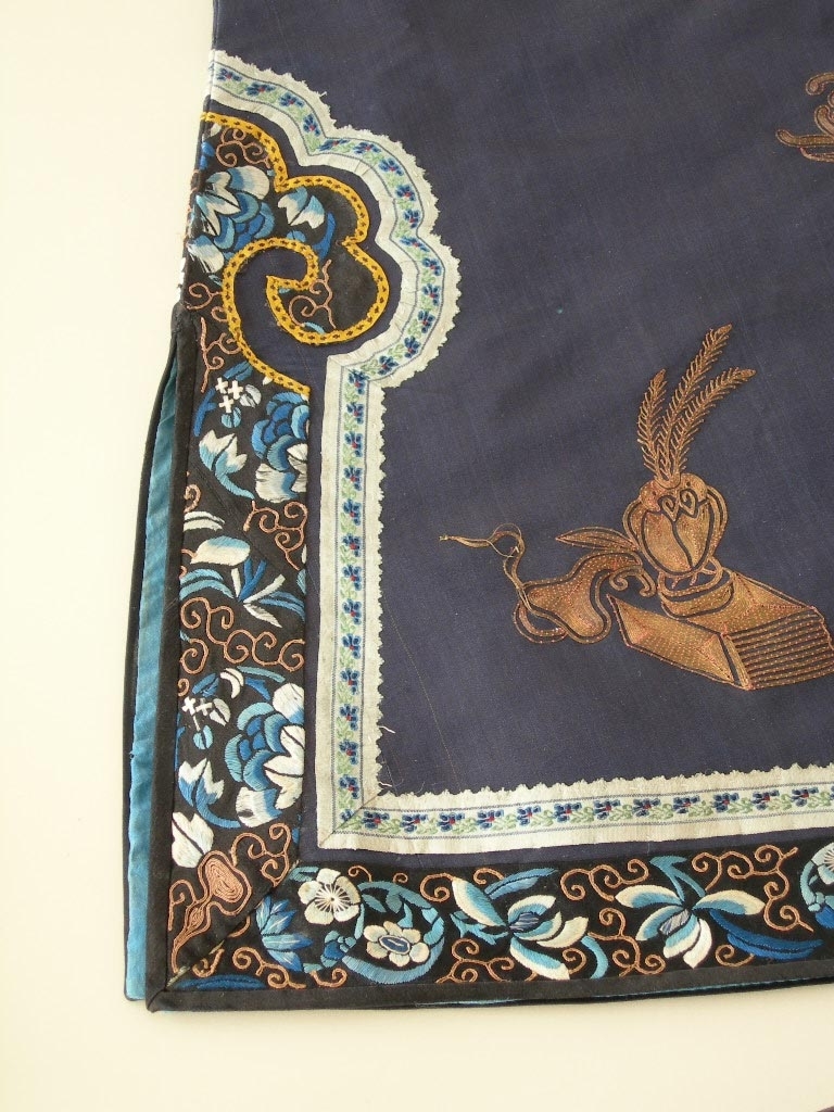 Kvinnojacka, troligen använd av mandarinhustru som ämbetsdräkt. Bärs vanligen tillsammans med byxor eller veckad kjol.

Blåsvart sidentyg, tuskaft. Rak ärm.
Fram- och bakstycke dekorerade med guldbroderi, läggsöm sydd med tvärstygn, roströd silketråd. Guldtråden är gjord av pappersguld virad runt en silkekärna. Vissa mönsterformer har en klassisk symbolik, t.ex. ärtskidan för fruktbarhet, andra former är av mer dekorativ art.

Dekorativa band runt framkant, nederkant och sprund. Ytterst ett kantband i blått siden, därefter ett band, silkebroderi, 3,5 cm, blommor och cirklar i vitt och olika blå nyanser samt guldbroderi, innerst ett mönstervävt band, 2 cm brett. I halsringningen ytterst ett kantband i blått siden, därefter ett band med silkebroderi, likadant som på kantningen runt nederkanten, därefter en snedremsa i siden, ca 1 cm bred, och innerst ett mönstervävt band, 1,5 cm brett.

På ärmen snedremsa i blått siden, ca 1,5 cm bred därefter band i silkebroderi likadant som runt nederkant och halsringning. Ytterst ett tyg, 12 cm brett, dekorerat med mönstervävt band i blått och guldfärg. Tyget är broderat? med tråd i liknande färg som tuskaftsbottnen. Åtdragen plattsöm som bildar mönster tillsammans med den något grövre tråden. Symboliska mönsterformer, de 8 taoistiska symbolerna, bl.a. flöjt, solfjäder och lotusblomma mellan dessa dekorativa mönster, S-formade. Ärmens baksida och ca 1/4 av framsidan täckt med guldbroderi, läggsöm sydd med tvärstygn, guldfärgad tråd, och detaljer i silkebroderi, svart, vitt och lila, platt- knut och klyvsöm. Mönstret föreställer bl.a. vattenströmmar, vågskvalp och en drake som stiger upp ur vattnet. Draken har 4 klor och symboliserar den näst högsta rangen, den högsta har 5 klor och det är kejsarens symbol. Draken är en positivt laddad symbol, en manlig symbol.

Jackan knäpps med tre klotformade mässingknappar och öglor gjorda av svarta, runda band.
Foder: ljusblått siden.

En märkning på höger framstyckes insida i halsringen, kinesiska tecken skrivet på tyget och på en vit klisterlapp.