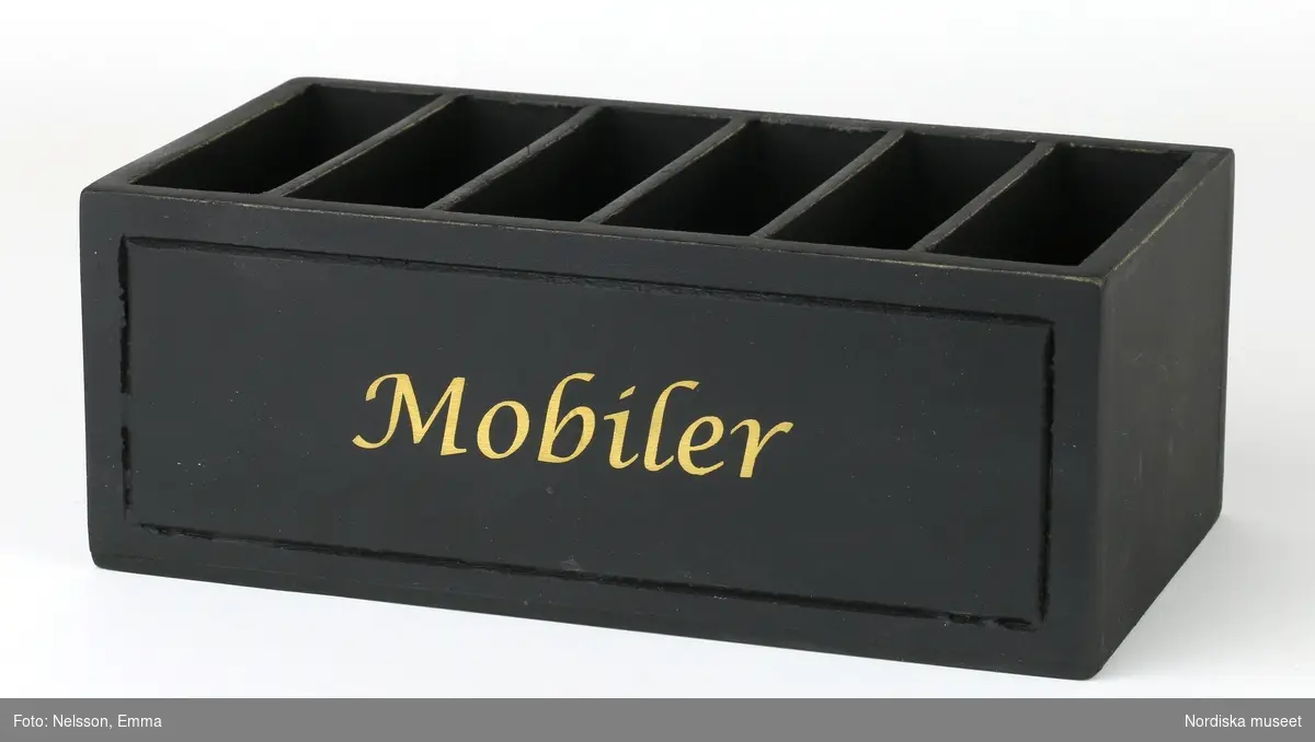 Rektangulär låda tillverkad i trä och målad i svart färg. Längs med långsidan återfinns texten "Mobiler" i guldfärg. Den innehåller förvaringsfack för sex mobiltelefoner.
/Anna Fredholm 2021-09-16