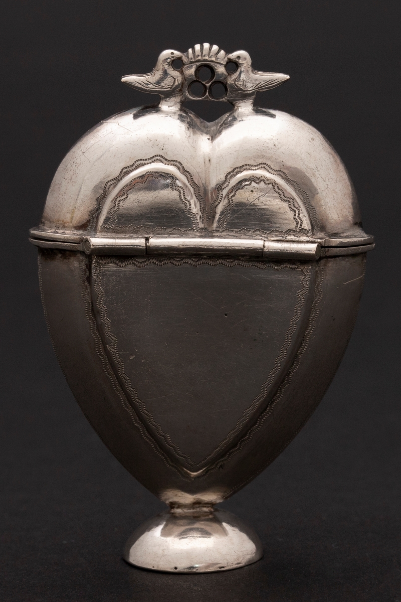 Hjerteformet luktevannshus i sølv med rund fot og to duer med kronelignede ornament mellom seg på lokket. Innvendig løs, perforert lokk. og svamp. Dekorert med prikkete bølgelinjer. Innvendig forgylt.