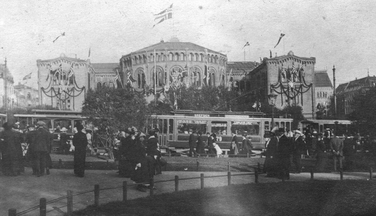 Sporvogner kjørende ved Eidsvolls plass 17.mai 1914, med mennesker i forgrunnen og Stortinget i bakgrunnen. 