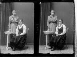 Portrett av to kvinner, en kvinne i bunad sitter foran ved e