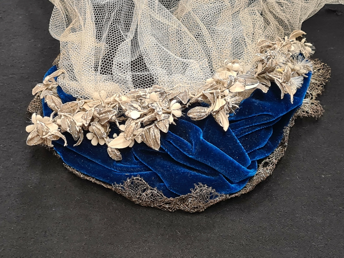 Hatt av blå sammet med metalltråd som dekor där fram och konstgjorda blommor i nacken. Till hatten, som är diademformad, är fäst en lång vit slöja i vit tyll.