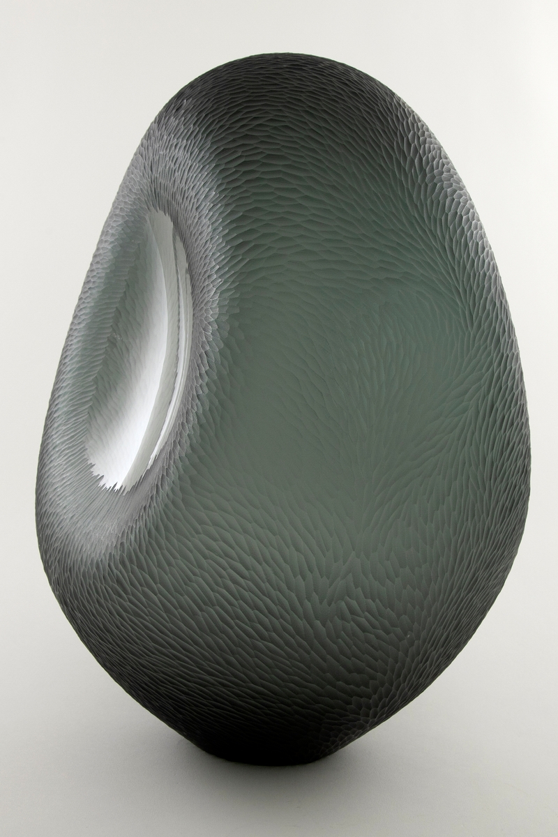Noe flattrykket ovalformet skulptur i gråfarget klart glass. Deler av overflaten er behandlet med sliping, sandblåsing og polering, slik at det oppstår en matt bølgende struktur over store deler av vasen. Midten av de konkave langsidene er ubehandlet, noe som muliggjør at betrakteren kan studere dekoren fra både ut- og innsiden. Sirkulær åpning i vasens bunn.