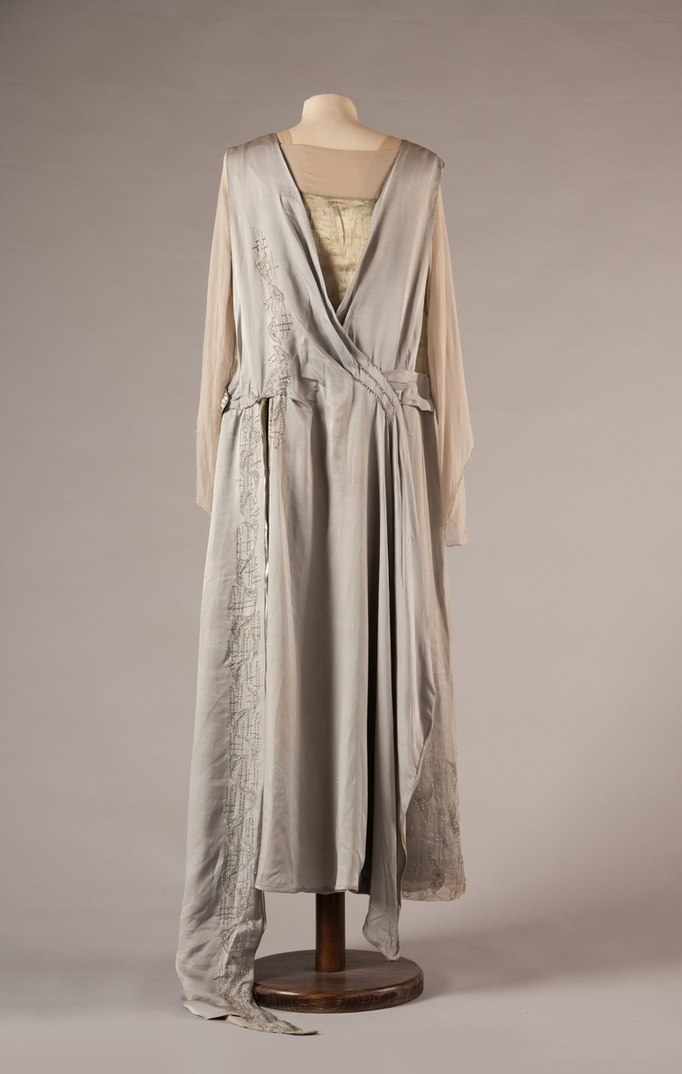 Selskapskjole i grått silkestoff med hvitt fôr i silkestoff. Kjolen er dekorert med glassperler (strass). Kjolen er produsert av Paulsson & Co i Oslo, trolig på 1920-tallet.