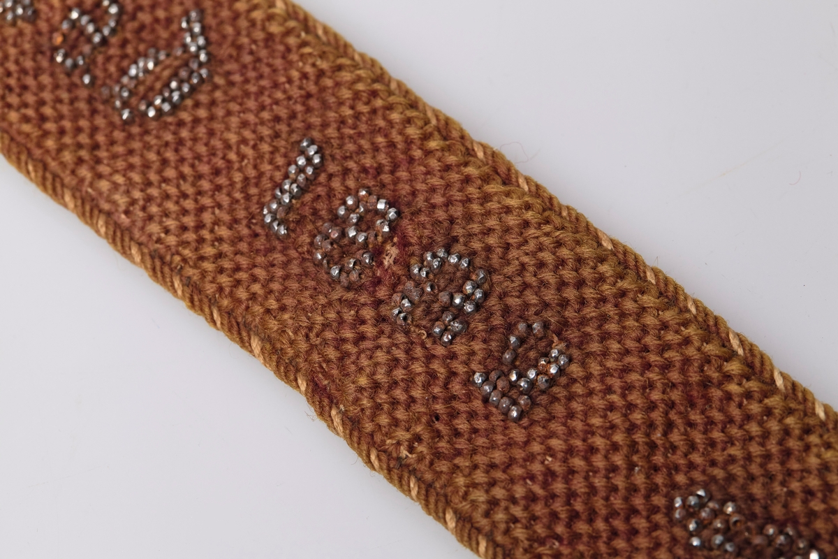 På den ene siden er det brodert "O. Rognstad 1892" med blanke perler på brun bunn. Motsatt side er dekorert med mange farger og har en knapp i hver ende.
