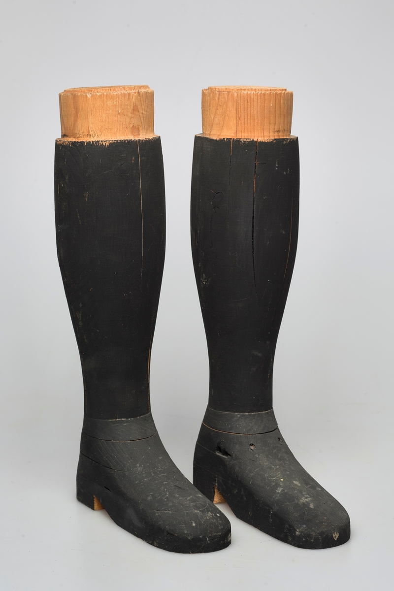 Svarte kompakte støvler av tre for utstilling. De er formet fullstendig etter kroppen og er del av en helfigur av en mann.