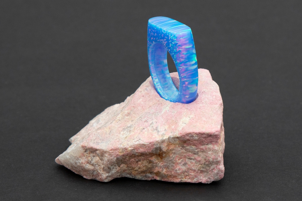 Lys blå ring med regnbue eller glittereffekter på rosa steinpodium. Ringen er lagd av syntetsik opal (bello) og podieet er av thulitt.