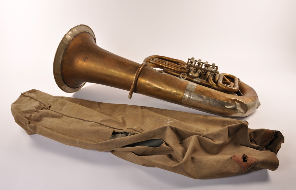 Tuba i messing, blankt metall, kork, lær. Mangler munnstykke. Tilhørende trekk i canvas med lærdetaljer (rifter, oppsprukket lær).