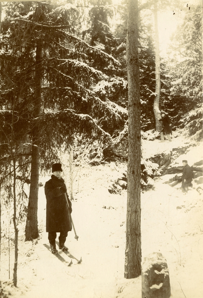 Henrich O.B.Homann på ski, en stav. En mann på ski på toppen av bakken og en til høyre på kne. I vinterskogen.