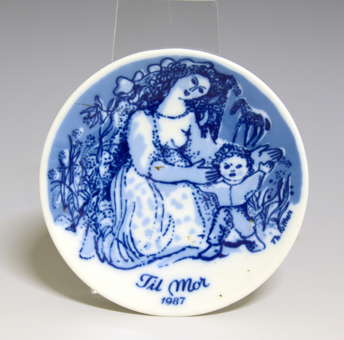 Morsdagsplatte av porselen. Hull til oppheng bak. Hvit glasur. Dekorert i blått med sittnde kvinne som støtter et lite barn, tekst Mors Dag 1987.