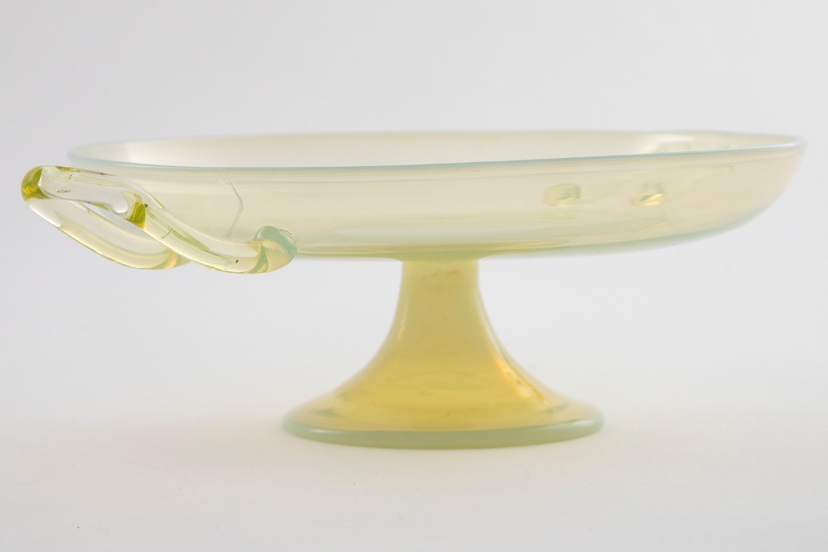 Skål i gultonet glass med opaliserende partier. Lav og vid kupa som bæres av en trumpetformet stett. Påsatte hanker på hver side av kupa.