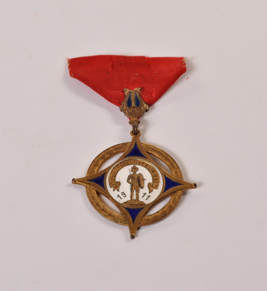 medalje i metall og emalje med rødt ordensbånd. Sangerstevnet i Fr.hald 1911